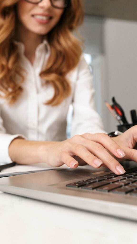 Woman in white blouse at work on laptop with notepad and pen nearby.