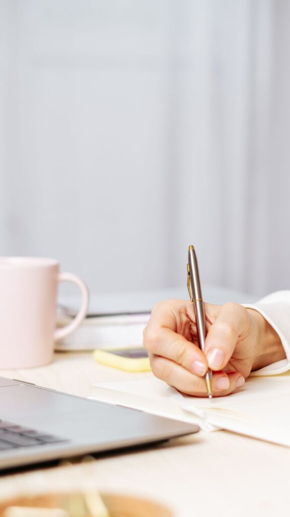 Woman writing in a notebook with a mug and laptop nearby.