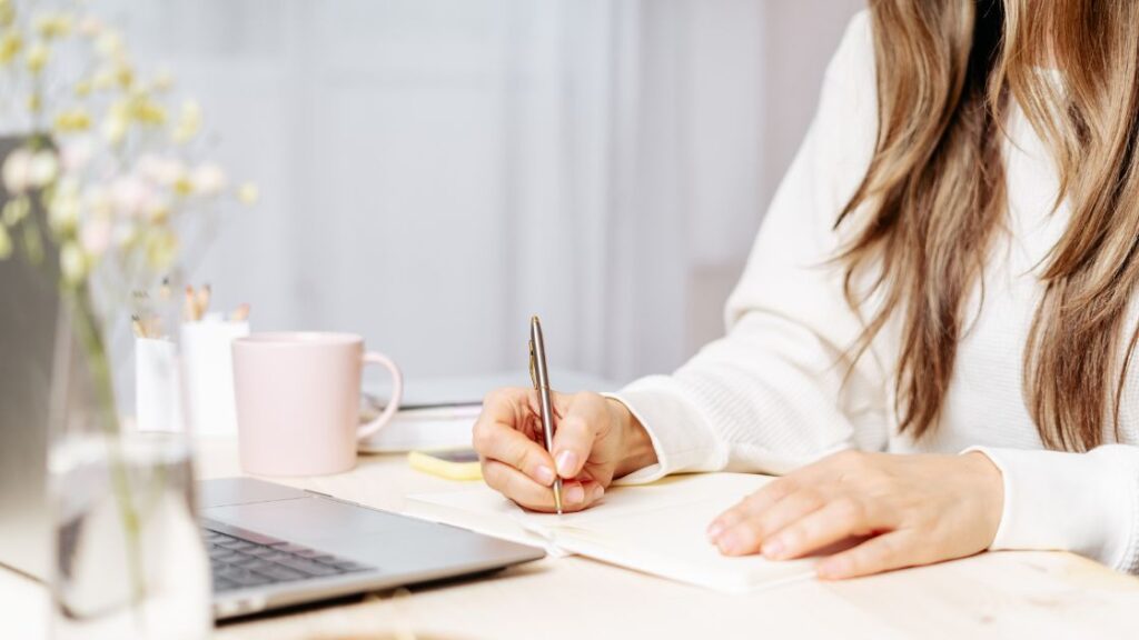 Woman writing in a notebook with a mug and laptop nearby.