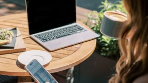 Laptop open on outdoor patio table. Woman seated nearby with cup of coffee in one hand and smartphone in the other.