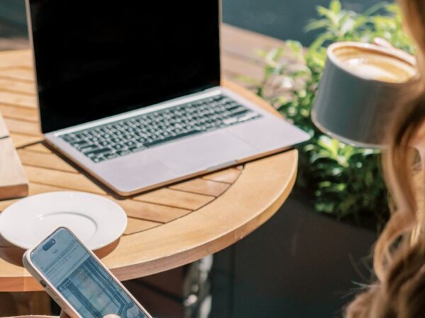 Laptop open on outdoor patio table. Woman seated nearby with cup of coffee in one hand and smartphone in the other.